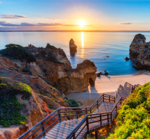 Holztreppe f&uuml;hrt zu einer versteckten Bucht mit Sandstrand und beeindruckenden Felsformationen an der Algarve bei Sonnenaufgang.