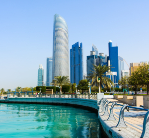Moderne Skyline von Abu Dhabi mit hohen Glasgeb&auml;uden am t&uuml;rkisfarbenen Wasser, ges&auml;umt von einer Promenade mit Palmen und Gel&auml;ndern.