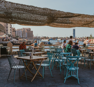 Stra&szlig;encaf&eacute; mit t&uuml;rkisfarbenen St&uuml;hlen am Dubai Creek, Menschen sitzen im Schatten mit Blick auf Boote und Skyline.