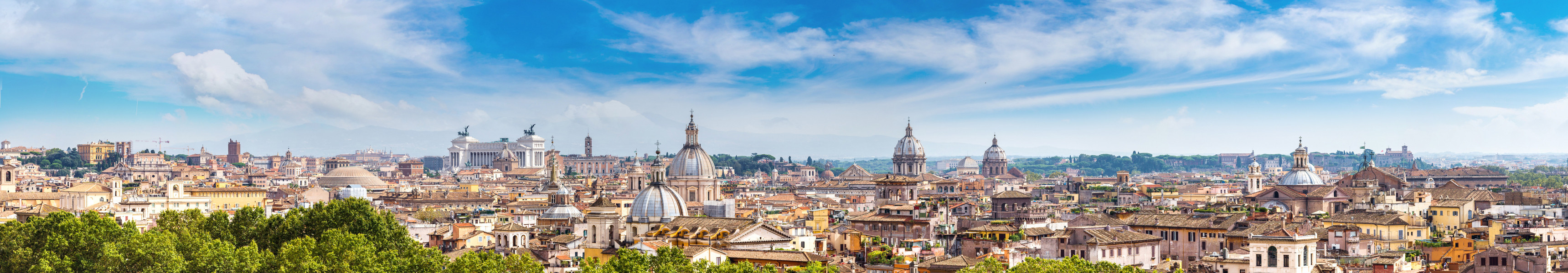 Panoramablick auf die D&auml;cher von Rom, mit markanten historischen Geb&auml;uden, Kirchen und Kuppeln, unter einem klaren blauen Himmel.