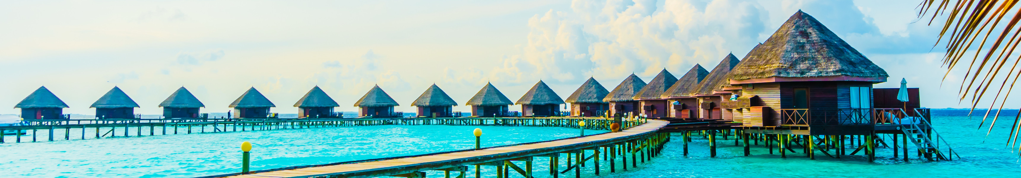 Reihen von &Uuml;berwasser-Bungalows mit Strohd&auml;chern auf den Malediven, verbunden durch Holzstege &uuml;ber t&uuml;rkisblauem Meer, eingerahmt von Palmenbl&auml;ttern im Vordergrund.