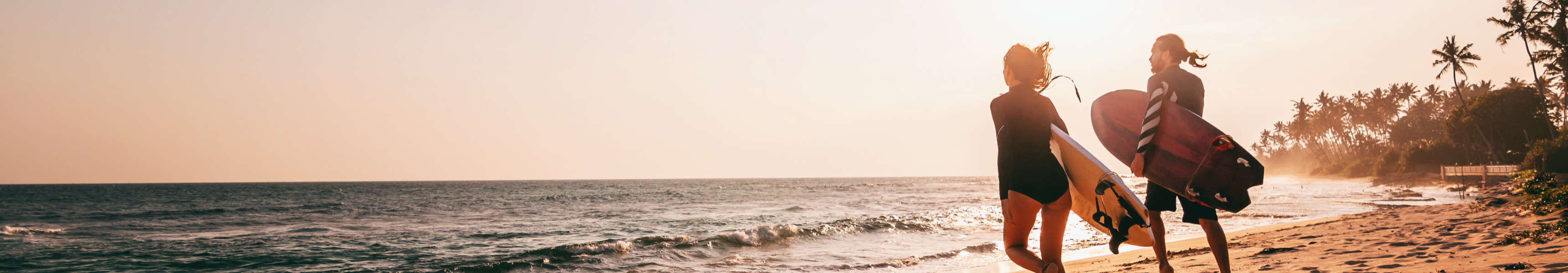 Zwei Surfer laufen mit ihren Brettern am Sandstrand entlang ins Meer, w&auml;hrend die Sonne &uuml;ber dem Ozean untergeht und Palmen die tropische Kulisse erg&auml;nzen.