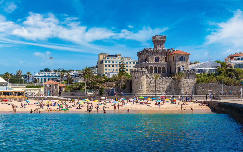 Sonniger Strand in Estoril mit goldenem Sand, t&uuml;rkisblauem Wasser und dem markanten Castelo dos Mouros im Hintergrund.