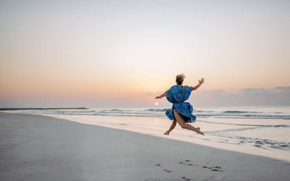 Junge Frau genie&szlig;t die Zeit am Strand und springt. Im Hintergrund sieht man das Meer mit Sonnenuntergang. 